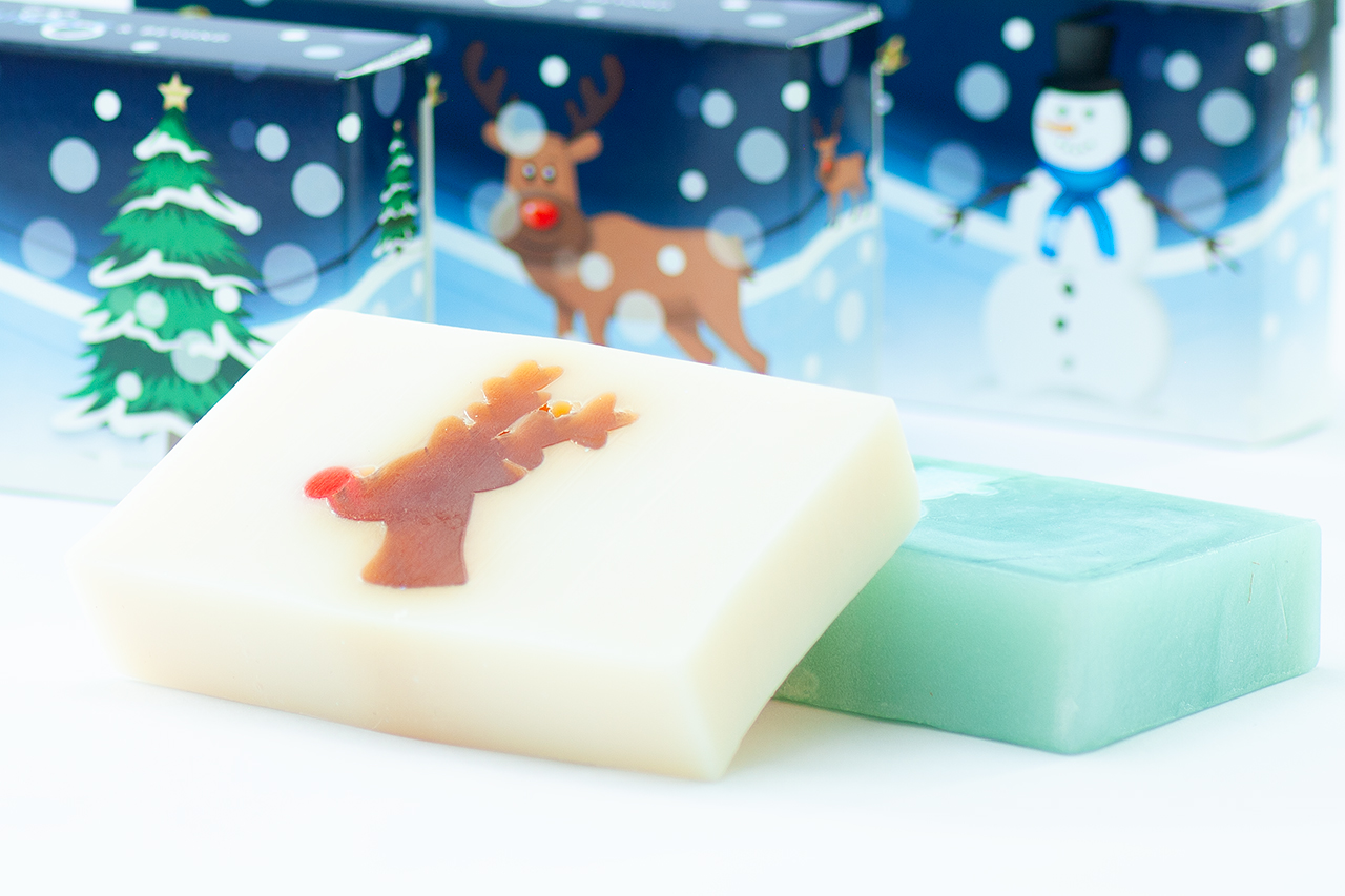 View of two festive soaps lying in front of three out of focus boxes featuring fun Christmas graphics.